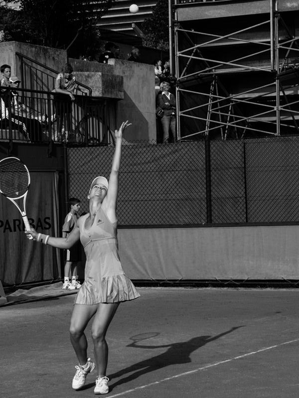 Female tennis player serving on outdoor court in black and white photography with spectators visible in background